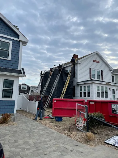 Crew on a house roof during a tear-off, with ladders and a red dumpster in the driveway